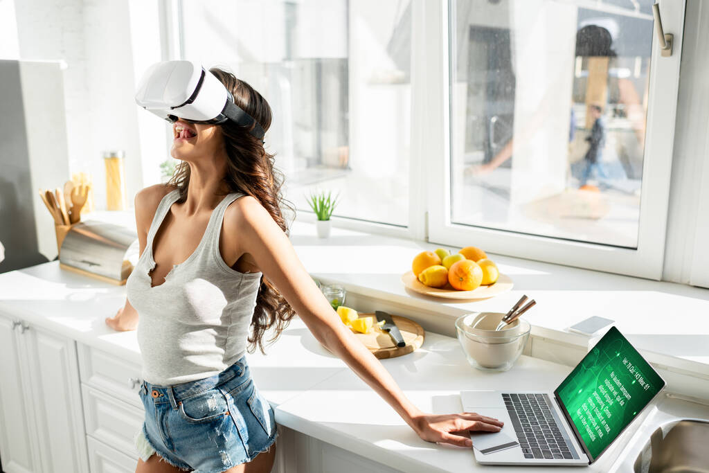 Женщина с помощью гарнитуры виртуальной реальности возле ноутбука и кредитной карты на кухне
  - Фото, изображение