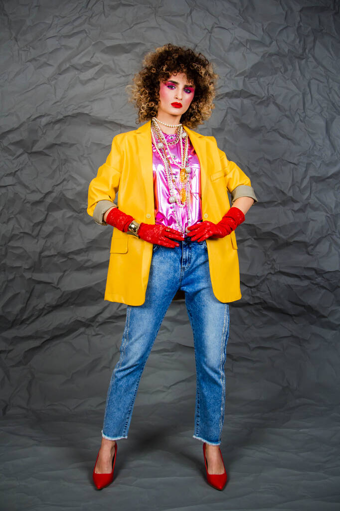 黄色のジャケットの女の子とアフロヘアスタイルの青いジーンズ。80年代のファッション、ディスコ時代。灰色の背景のスタジオ写真. - 写真・画像