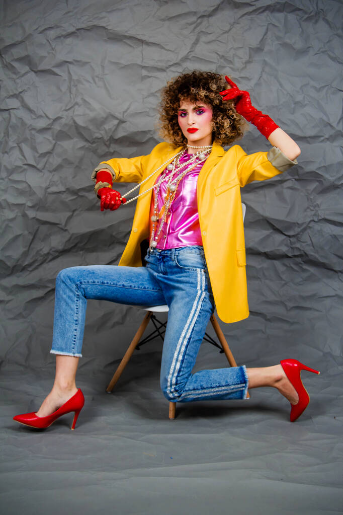 A girl in a yellow jacket and blue jeans with an afro hairstyle sits on a chair. Fashion eighties, the era of disco. Studio photo on a gray background. - Photo, Image