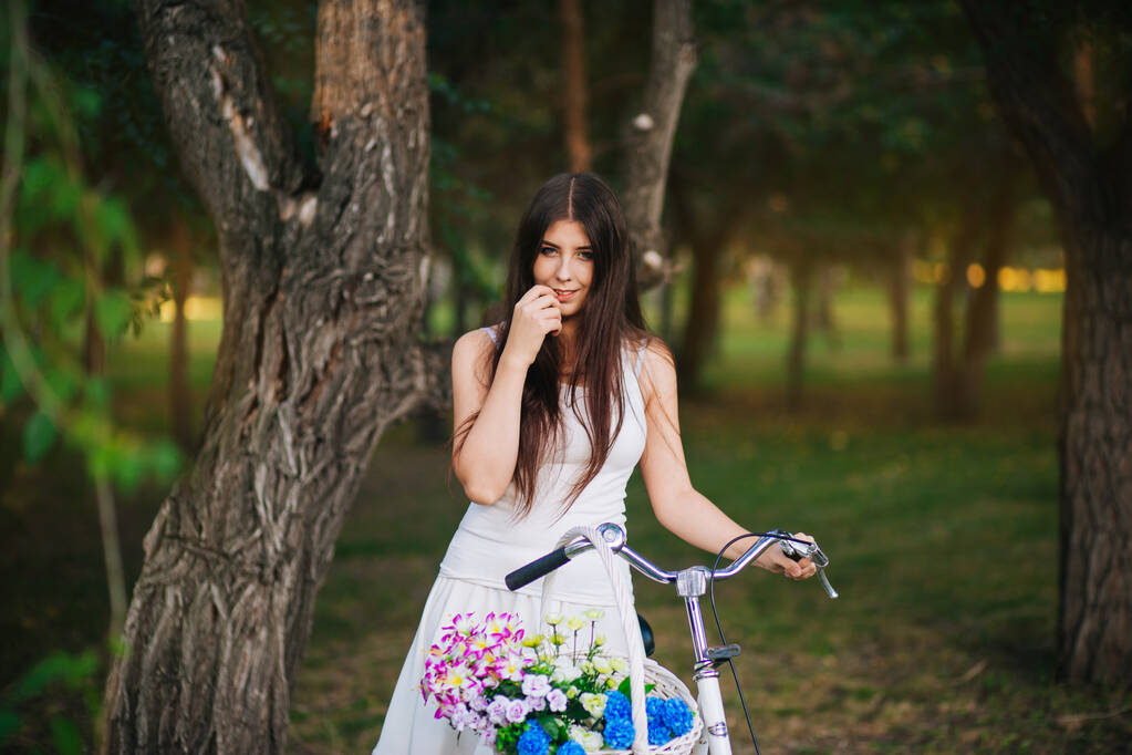 Retrato de una niña en una camiseta blanca y falda con una bicicleta y una cesta de flores en el parque por la noche
. - Foto, Imagen