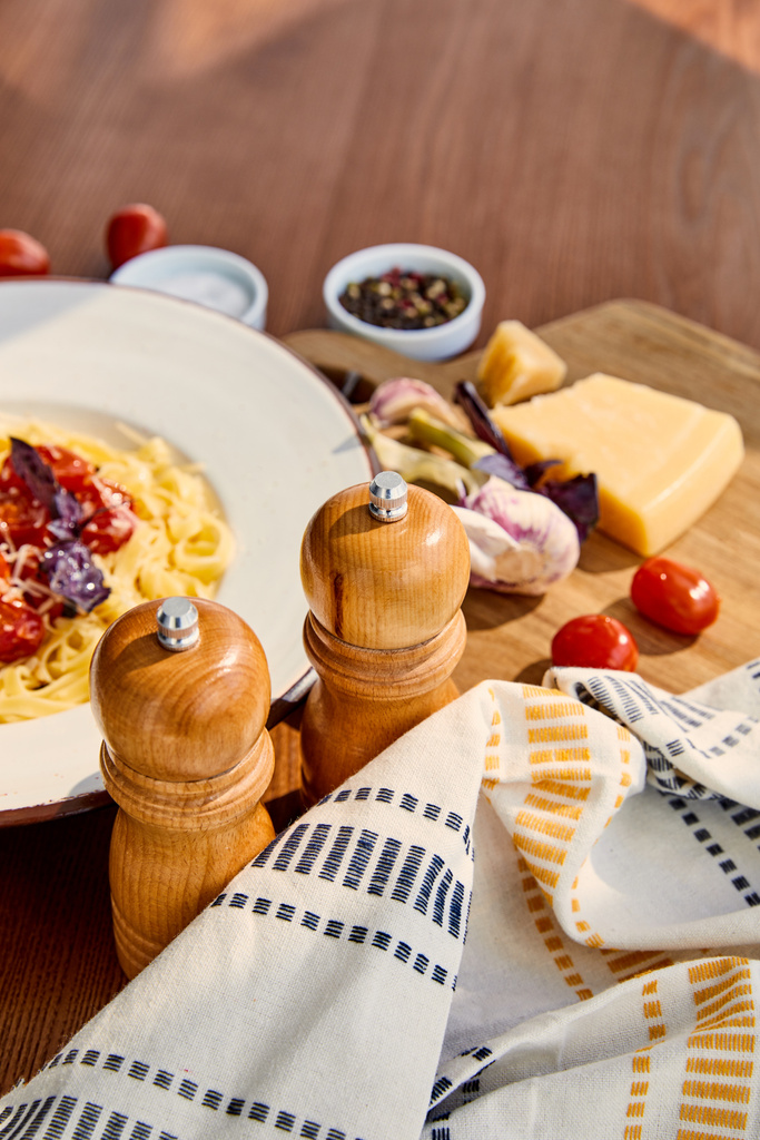 selective focus of delicious pasta with tomatoes served on wooden table with cutlery, napkin, seasoning and ingredients in sunlight - Photo, Image