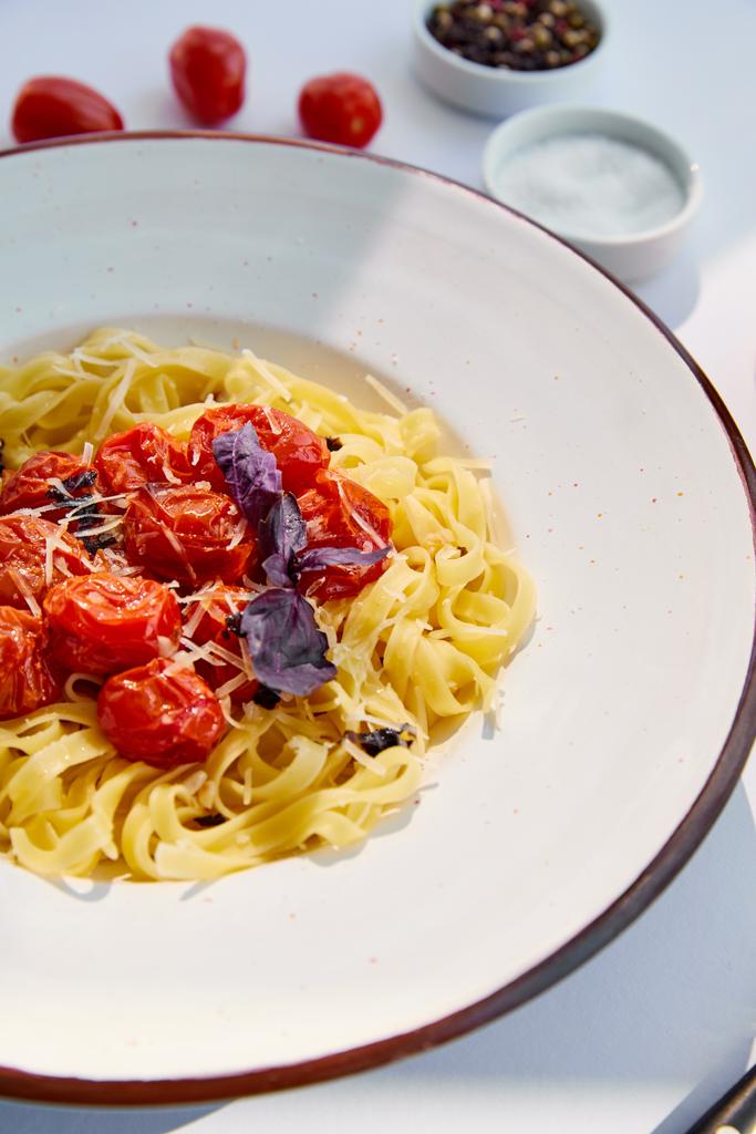 enfoque selectivo de deliciosa pasta con tomates servidos con sal y pimienta en la mesa blanca a la luz del sol
 - Foto, Imagen