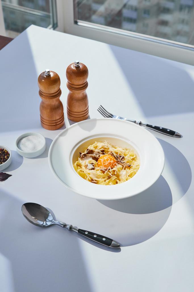 вкусная паста карбонара подается с столовыми приборами, соль и перец мельницы на белом столе в солнечном свете
 - Фото, изображение