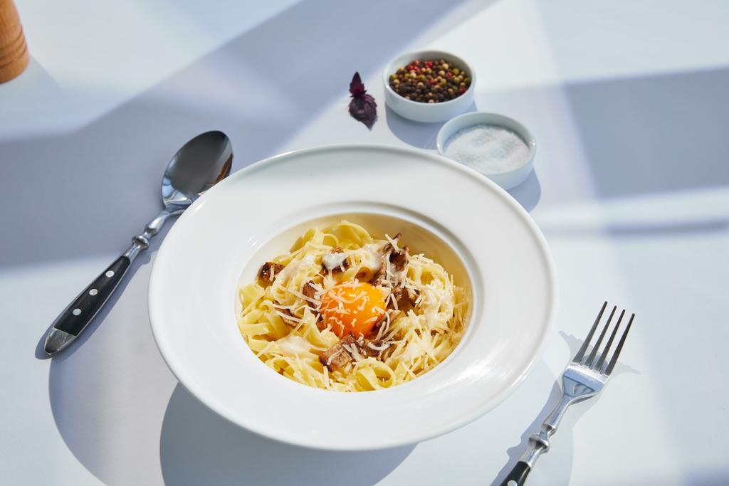 heerlijke pasta carbonara geserveerd met bestek en kruiden op witte tafel in zonlicht - Foto, afbeelding