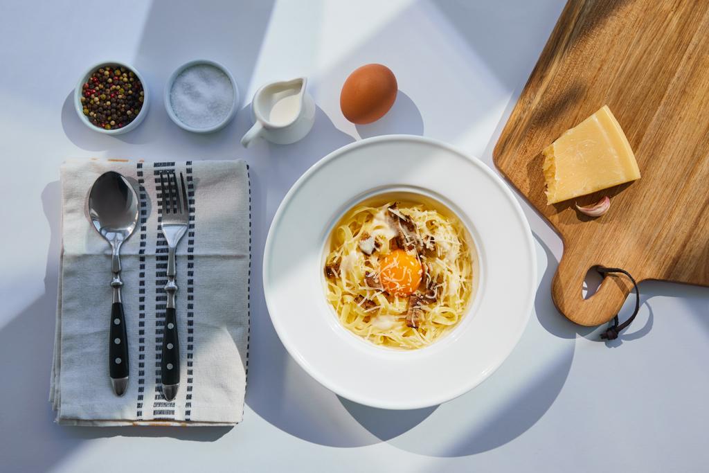 ylhäältä näkymä herkullinen pasta carbonara tarjoillaan ruokailuvälineet, mausteet ja ainesosat valkoisella pöydällä auringonvalossa
 - Valokuva, kuva