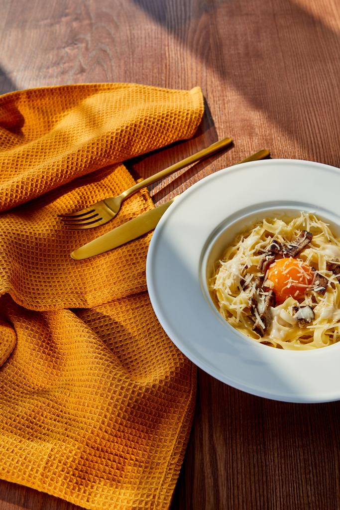 köstliche Pasta Carbonara serviert mit goldenem Besteck und gelber Serviette auf Holztisch im Sonnenlicht - Foto, Bild