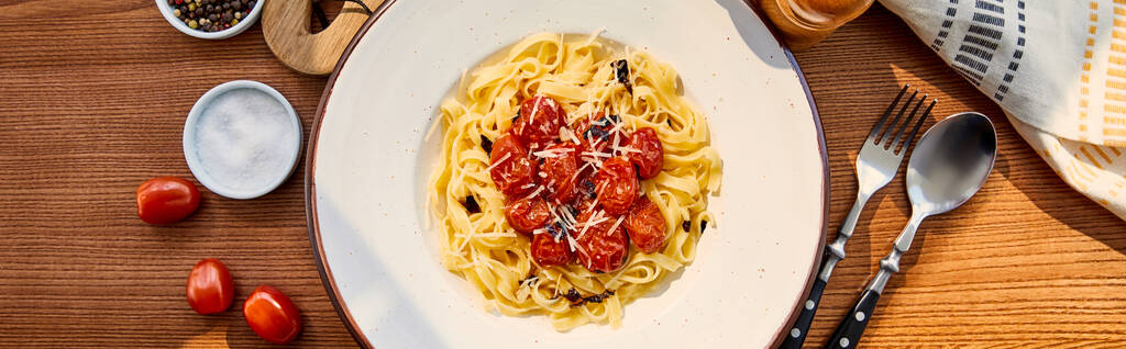 top view of delicious pasta with tomatoes served on wooden table with cutlery, napkin, seasoning and napkin in sunlight, panoramic shot - Photo, Image