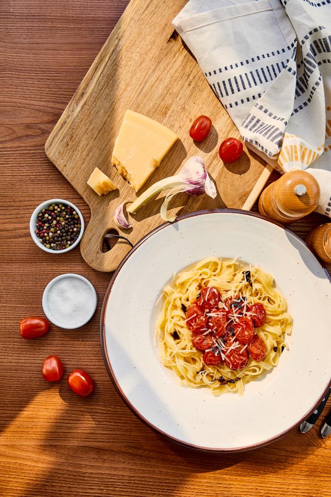 top view of delicious pasta with tomatoes served on wooden table with cutlery, napkin, seasoning and ingredients in sunlight - Photo, Image