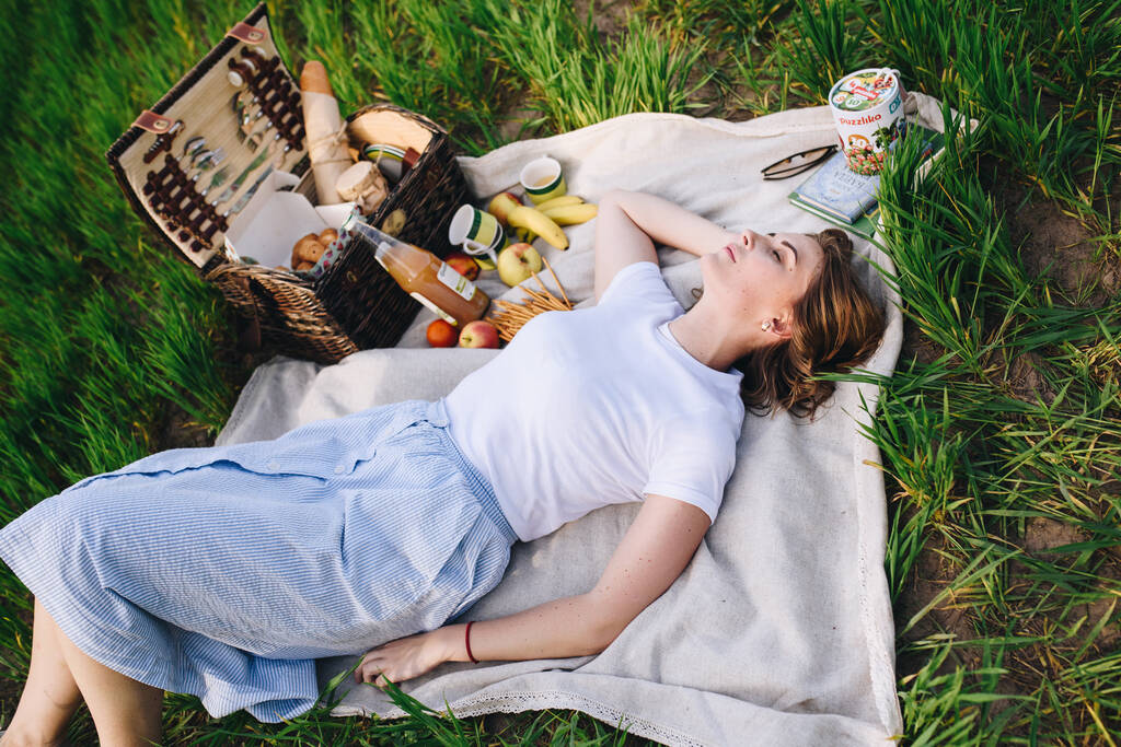 Una chica en el bosque en un picnic. Se sienta en un claro, hierba verde. Ropa azul. Cesta de picnic con comida. Almuerzo en la naturaleza. Rubia, pelo corto. El lavado a la moda
. - Foto, imagen