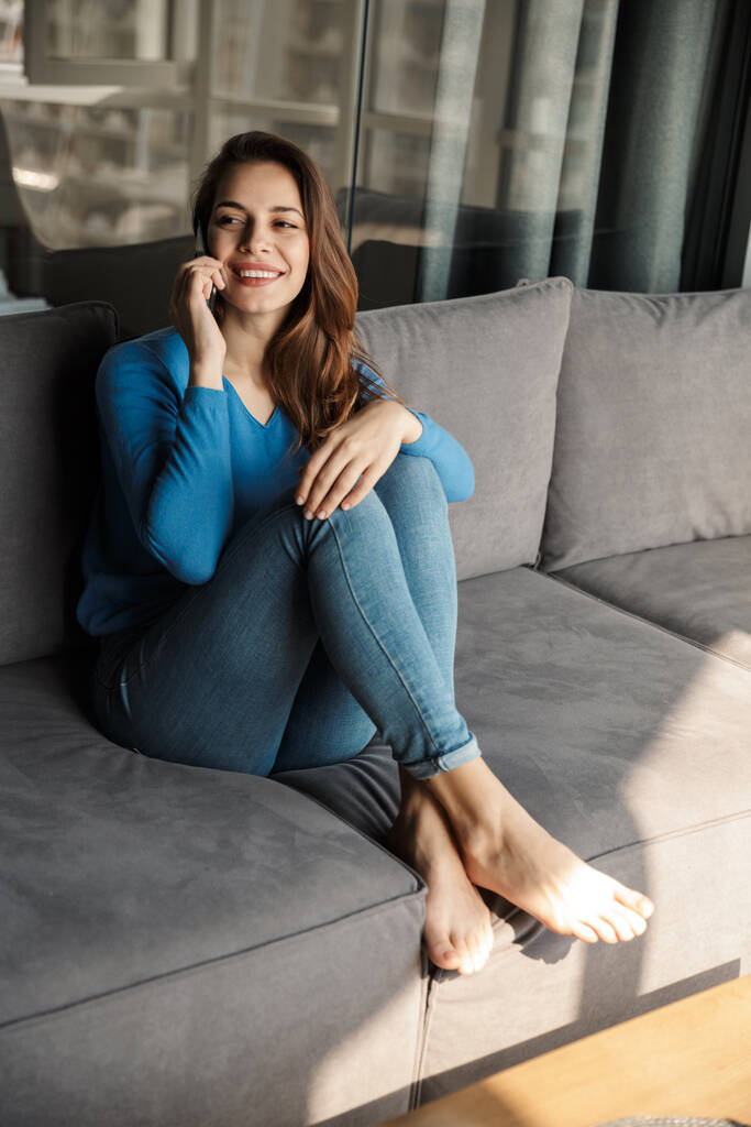 Bild von netter junger zufriedener Frau, die auf dem Sofa im Wohnzimmer sitzt und mit dem Handy spricht und lächelt - Foto, Bild