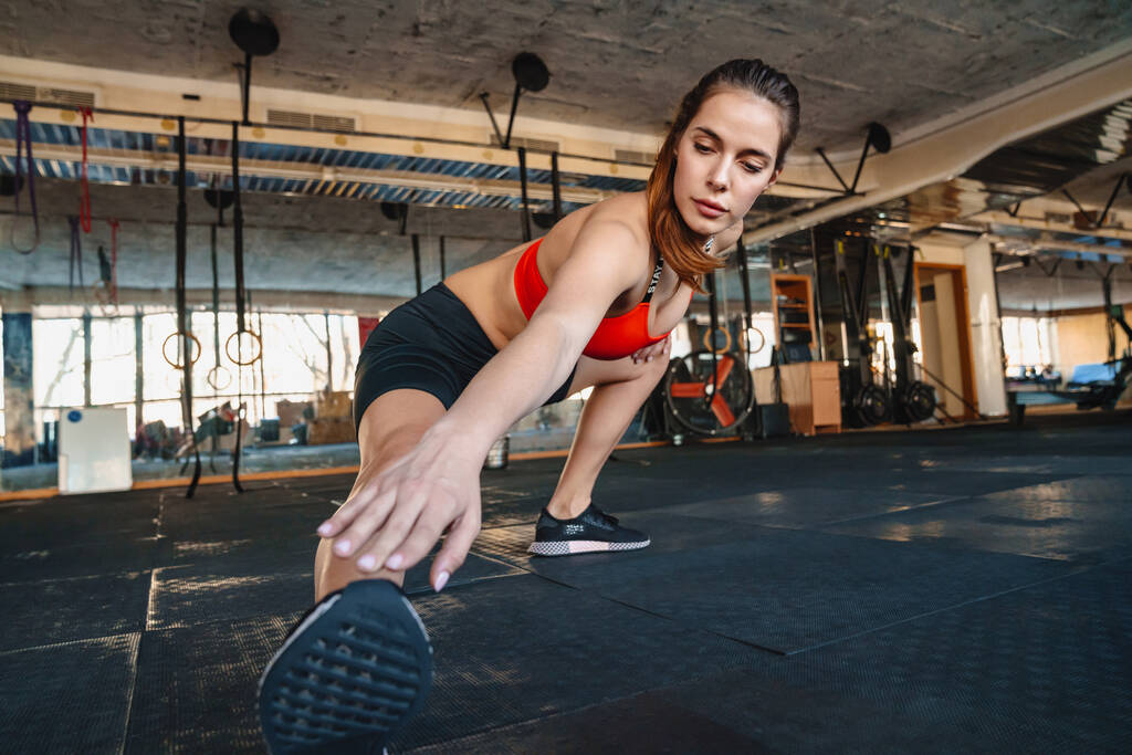 Portrét atletické krásné sportovkyně protahuje nohu při cvičení v atletické hale - Fotografie, Obrázek