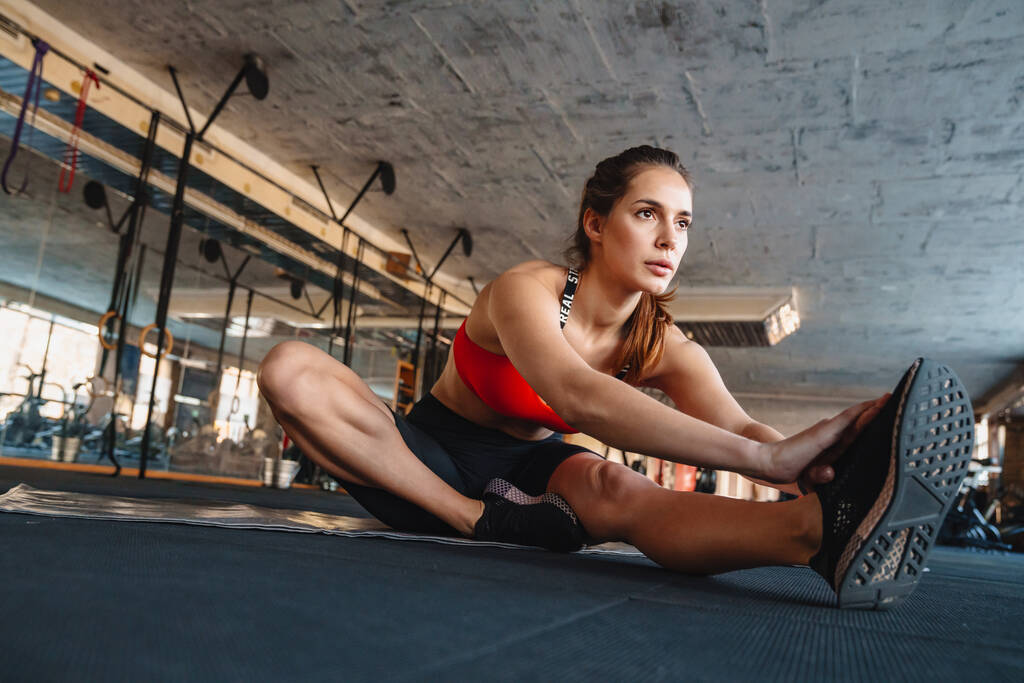 Портрет спортивной красивой спортсменки, растягивающей ногу во время тренировки в спортзале
 - Фото, изображение