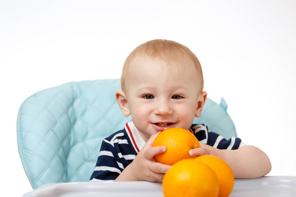 Iloinen poika istuu lastenistuimella ja syö appelsiineja valkoisella taustalla. Tuoreet hedelmät ovat erittäin hyödyllisiä lapsille. Älykkäät aikuiset ovat hyviä ravitsemusterapeutteja
. - Valokuva, kuva
