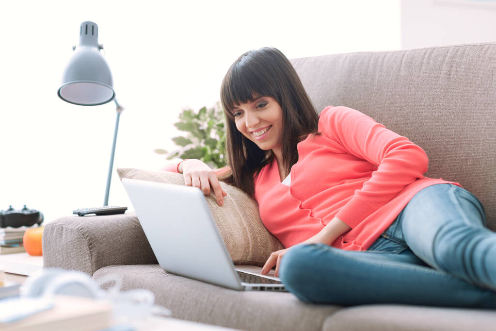 Evde gülümseyen genç bir kadın, kanepede rahatlıyor ve bir dizüstü bilgisayarla sosyal iletişim kuruyor. - Fotoğraf, Görsel
