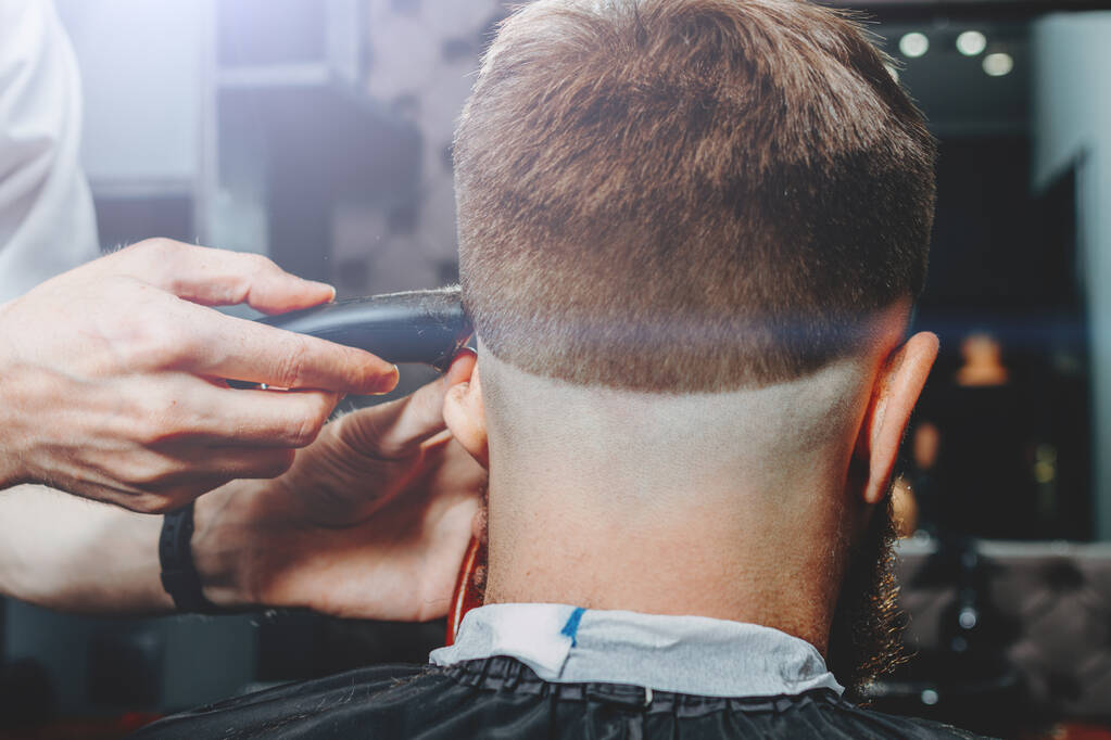 BARBERSHOP THEME. BARBER IS TRIMMING THE HAIRCUT OF HIS BEARDED CLIENT. HE IS USING A HAIR CLIPPER - Photo, Image