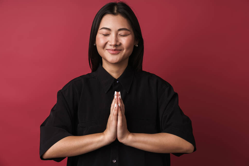 Zdjęcie azjatyckiej optymistycznej, relaksującej młodej kobiety pozującej na czerwonym tle muru, wykonującej gest medytacji.. - Zdjęcie, obraz