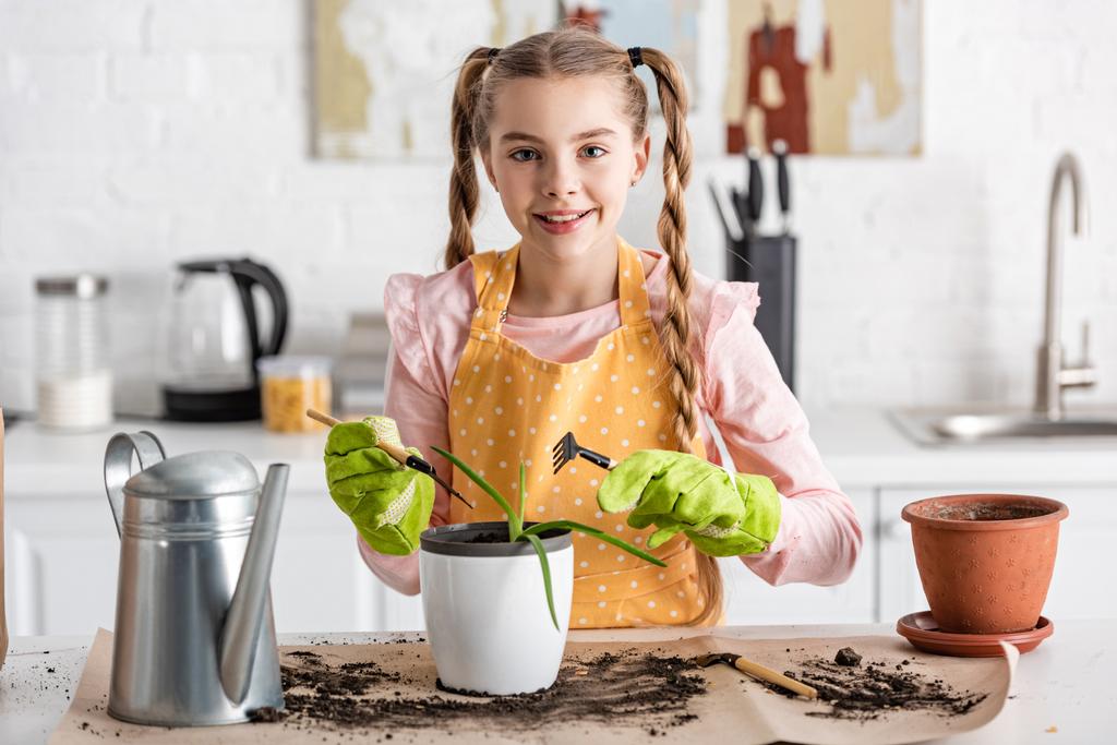 Vooraanzicht van schattig kind met tuingereedschap glimlachend bij tafel met drinkpot, grond- en bloempotten in keuken - Foto, afbeelding