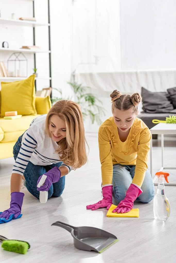 Mother and daughter with spray bottles and rags wiping floor near brush and dustpan in living room - Photo, Image