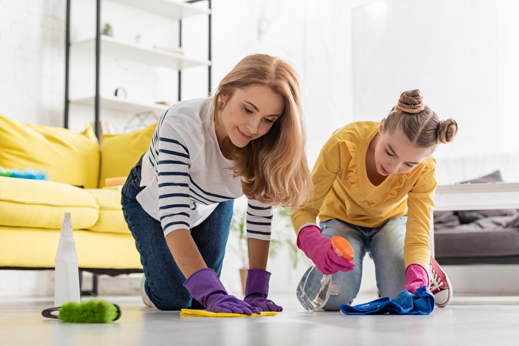 Mother and daughter with spray bottle and rags wiping floor near brush in living room - Photo, Image