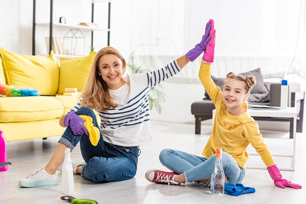 Madre e hija con artículos de limpieza dando choco cinco, sonriendo y mirando a la cámara en el suelo en la sala de estar
 - Foto, Imagen