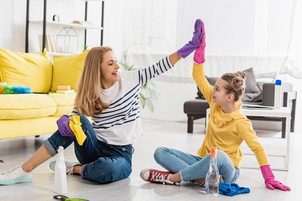 Mãe e filha com suprimentos de limpeza dando alta cinco, sorrindo e olhando um para o outro no chão na sala de estar
 - Foto, Imagem