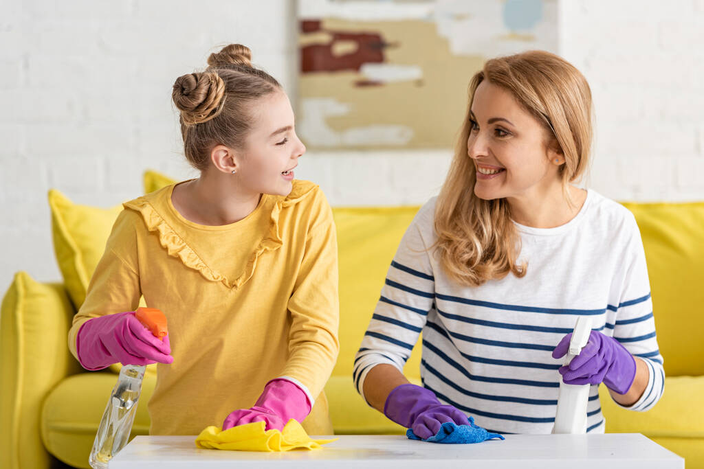 Mother and daughter with spray bottles smiling, looking at each other and wiping coffee table with rags near sofa in living room - Photo, Image