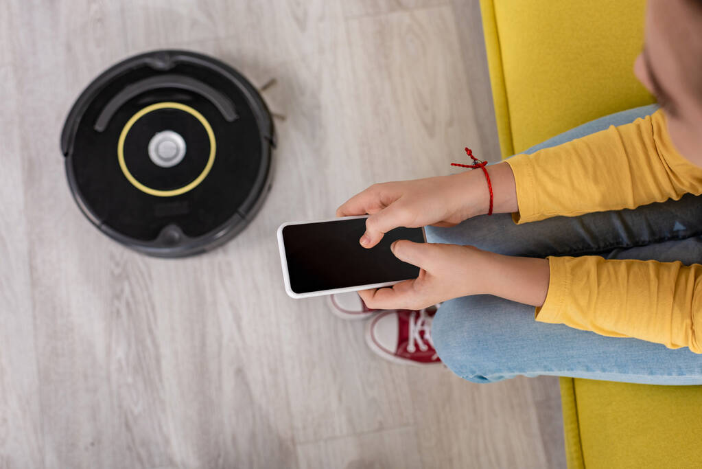Ausgeschnittene Ansicht von Kind mit Smartphone auf Sofa in der Nähe von Staubsaugerroboter auf dem Boden im Wohnzimmer - Foto, Bild