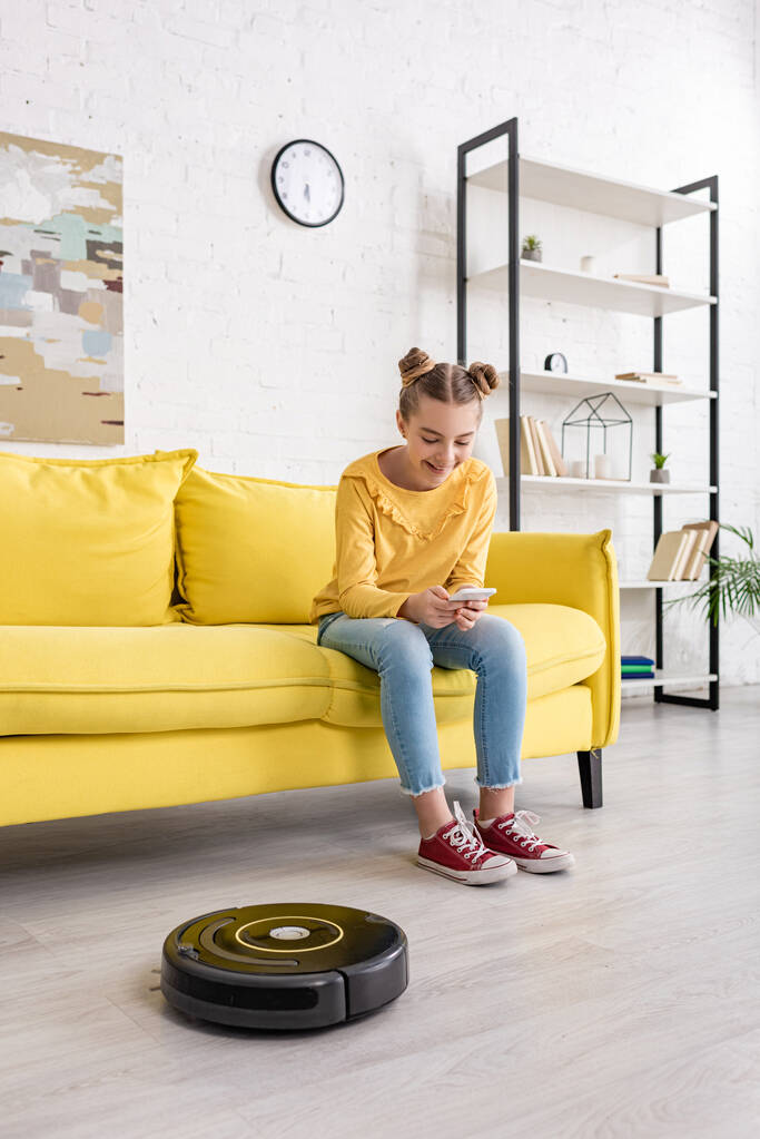 Cute child with smartphone smiling on sofa near robotic vacuum cleaner on floor in living room - Photo, Image