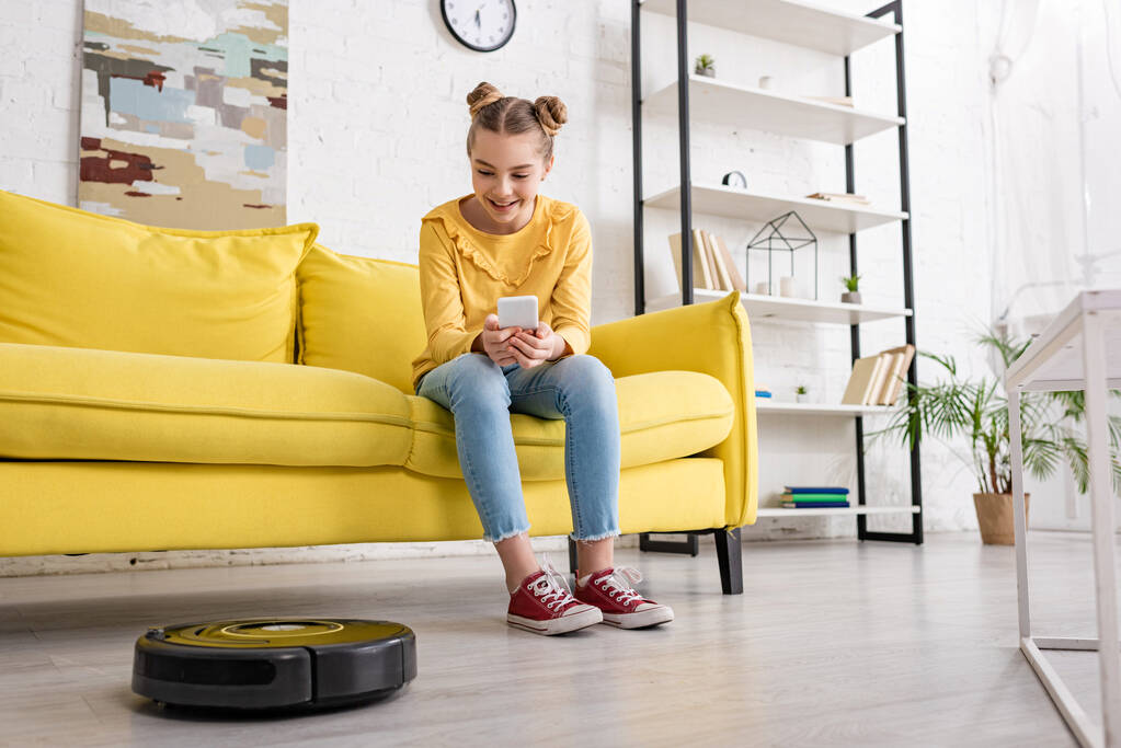 ソファの上でスマートフォンを持つかわいい子供は笑顔で、リビングルームの床にロボット掃除機を見て - 写真・画像