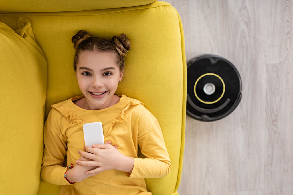 Top view of cute child with smartphone smiling, looking at camera and lying on sofa near robotic vacuum cleaner on floor in living room - Photo, Image