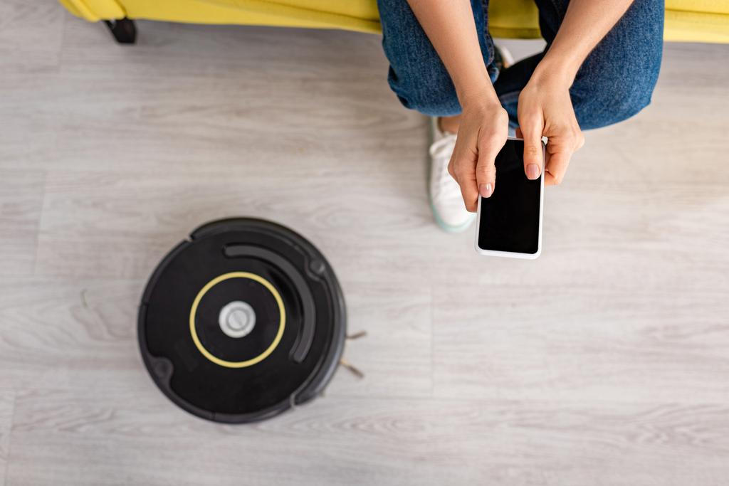 Ausgeschnittene Ansicht einer Frau, die ihr Smartphone in der Nähe eines Staubsaugerroboters auf dem Boden im Wohnzimmer hält - Foto, Bild