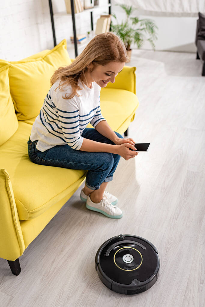 High angle view of woman smiling with smartphone on sofa near robotic vacuum cleaner on floor in living room - Photo, Image
