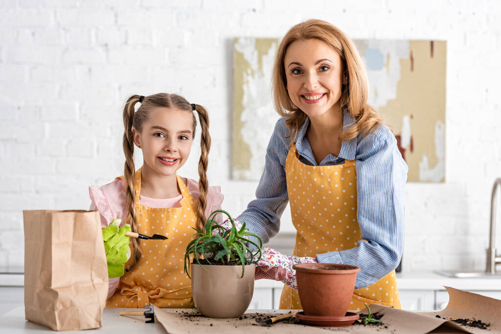 Mãe feliz e filha com ferramentas de jardinagem, vasos com aloés e saco de papel com chão sorrindo perto da mesa na cozinha
 - Foto, Imagem