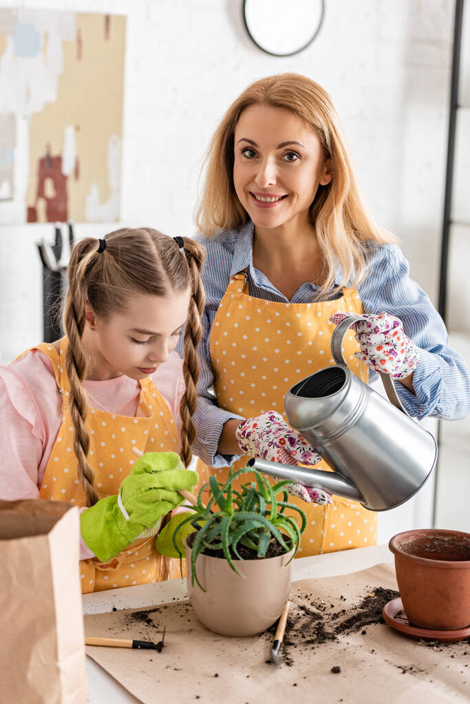 Vrouw lachend met dochter en met waterpot boven bloempot met aloë bij tafel in keuken - Foto, afbeelding