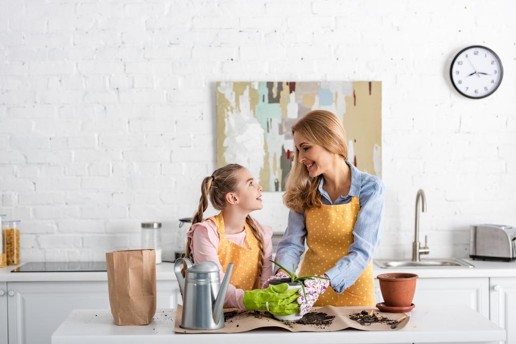 Mother and cute daughter looking at each other and touching flowerpot with aloe near watering pot and paper bag on table with ground - Photo, Image