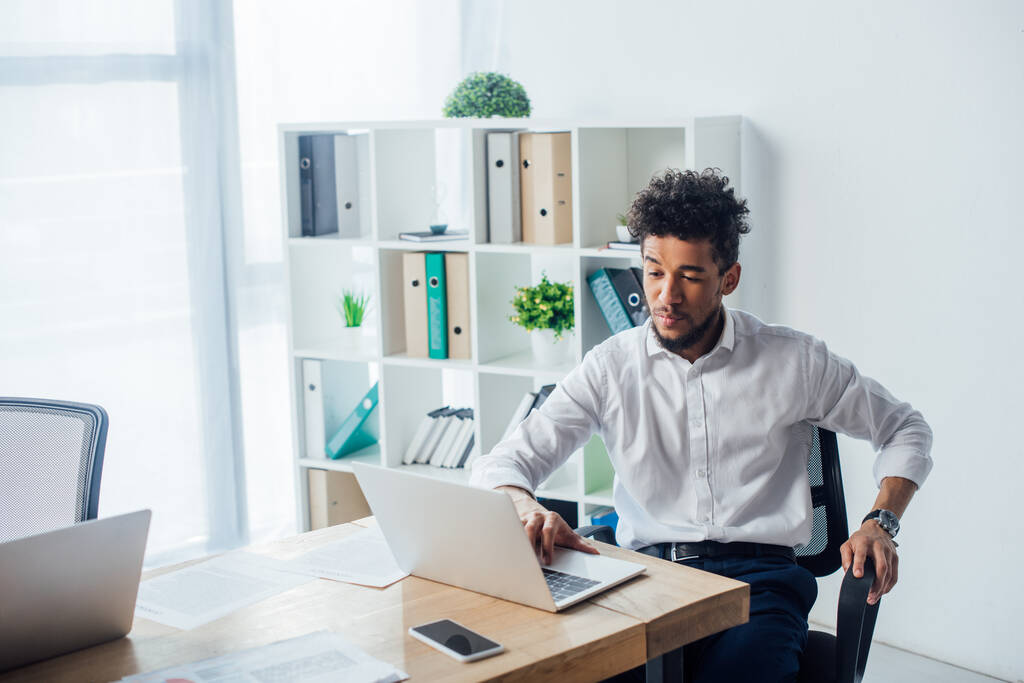 Afrikkalainen amerikkalainen liikemies käyttää kannettavaa tietokonetta lähellä papereita pöydällä
  - Valokuva, kuva