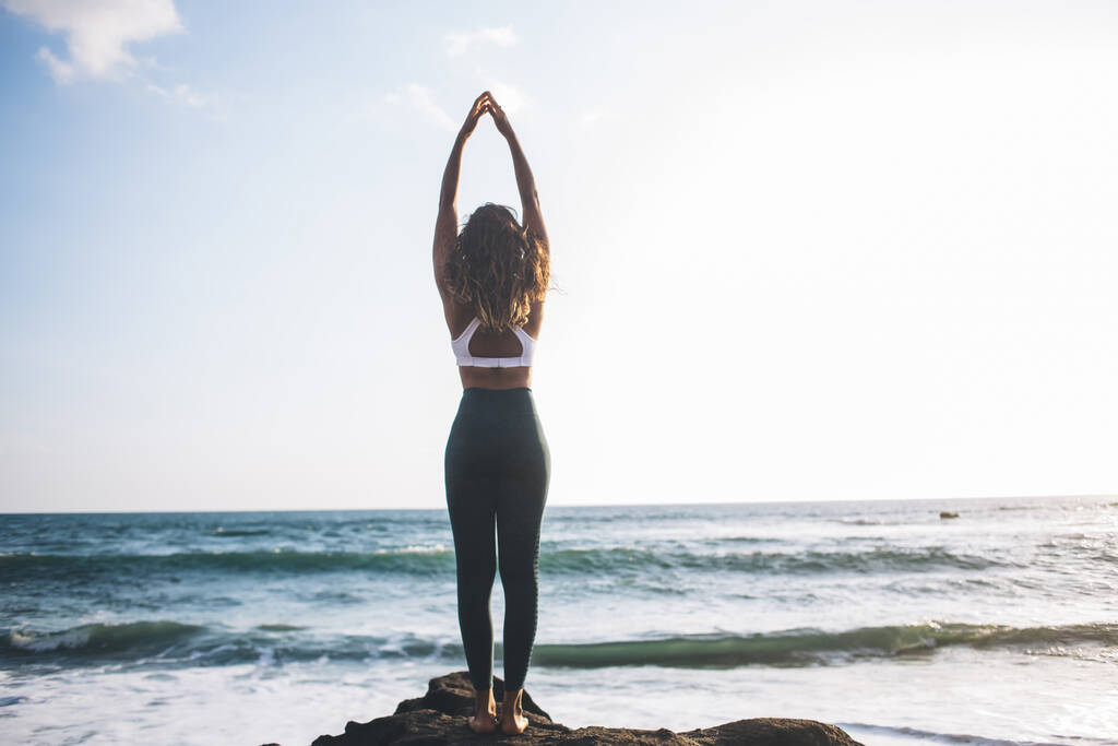 Achteraanzicht van jonge vrouw verhoogt lichaamsbewustzijn terwijl het doen van tadasana pose in de buurt van de oceaankust genieten van tijd voor het recreëren ziel, concept van gezonde levensstijl en harmonie met yoga meditatie - Foto, afbeelding