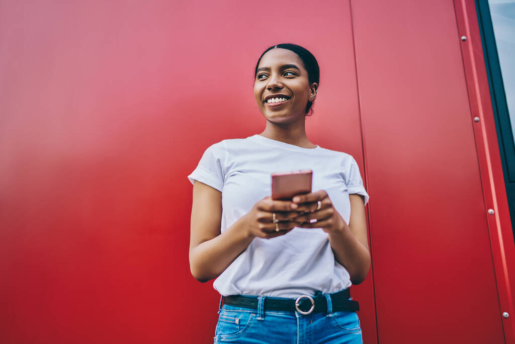 若いアフリカ系アメリカ人女性を笑顔でシャツにコピースペース用ブランド名またはラベル保持携帯電話と離れて見て、明るい暗い肌の女性ブロガー使用してスマートフォンスタンドon赤壁 - 写真・画像