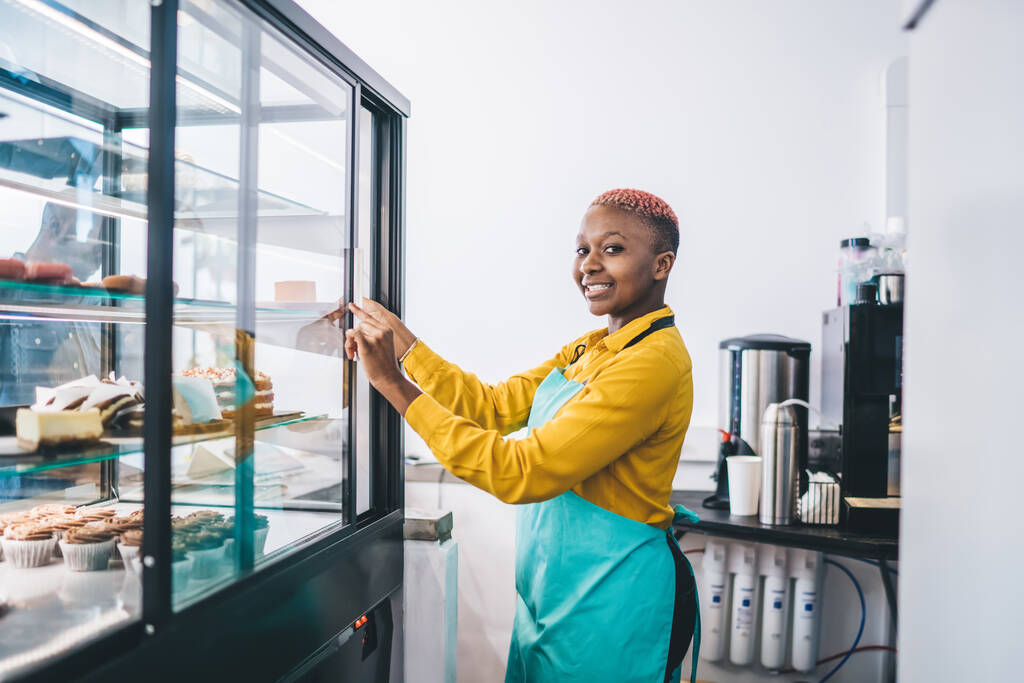 Vue latérale de la femme afro-américaine joyeuse souriante et vitrine d'ouverture avec des desserts frais tout en travaillant dans une petite boulangerie
 - Photo, image