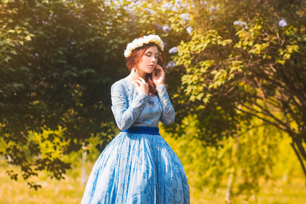 Atractiva mujer en un magnífico vestido azul camina en el parque sobre el fondo de flores de color lila en flor
. - Foto, Imagen