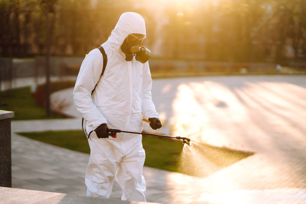 特別な保護消毒スーツを着た男は、隔離の街の夜明けに空の公共の場所で滅菌器を噴霧します。COVID -19 。清掃コンセプト. - 写真・画像