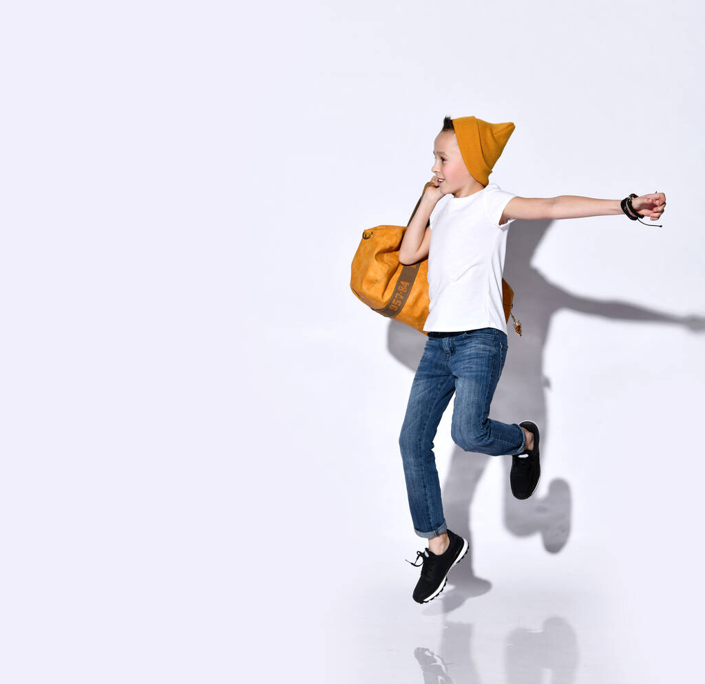 Adolescent avec sac de voyage orange, chapeau, t-shirt, jean bleu, baskets noires. Il saute, sourit, pose isolé sur blanc
 - Photo, image