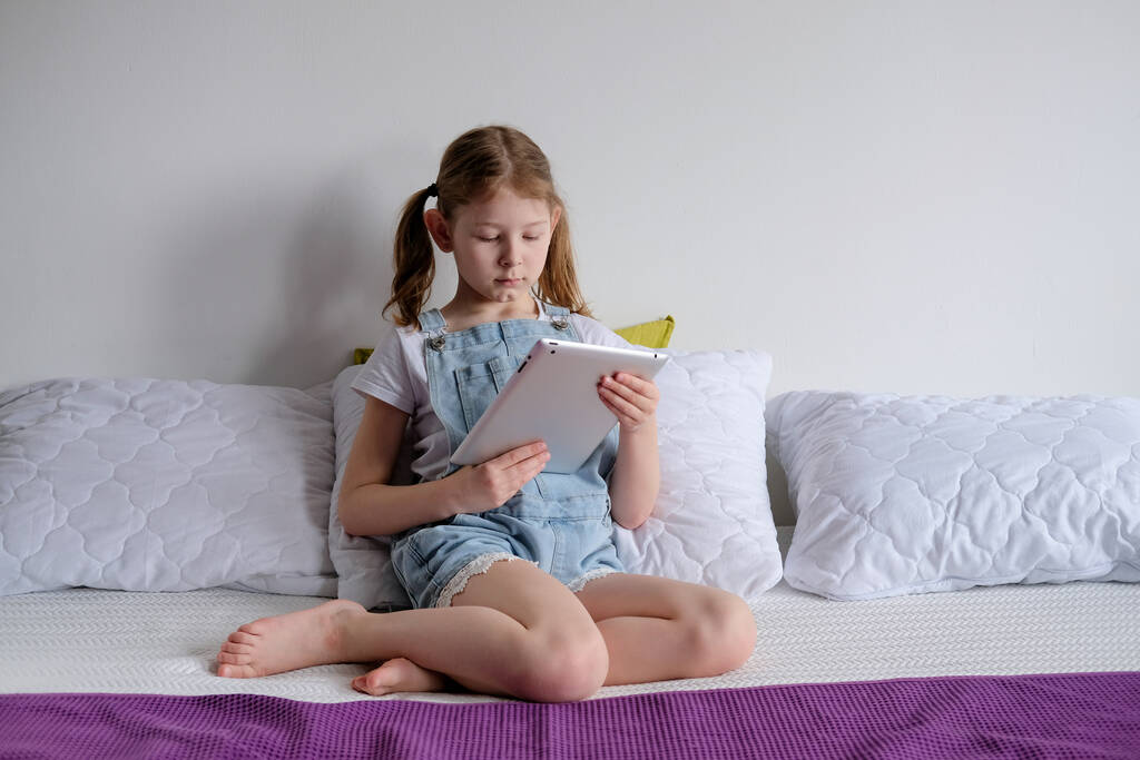 A lány otthon ül az ágyon, a kezében pedig egy laptop. Egy iskoláslány tanul és játszik egy laptopon. Karantén önképzés - Fotó, kép