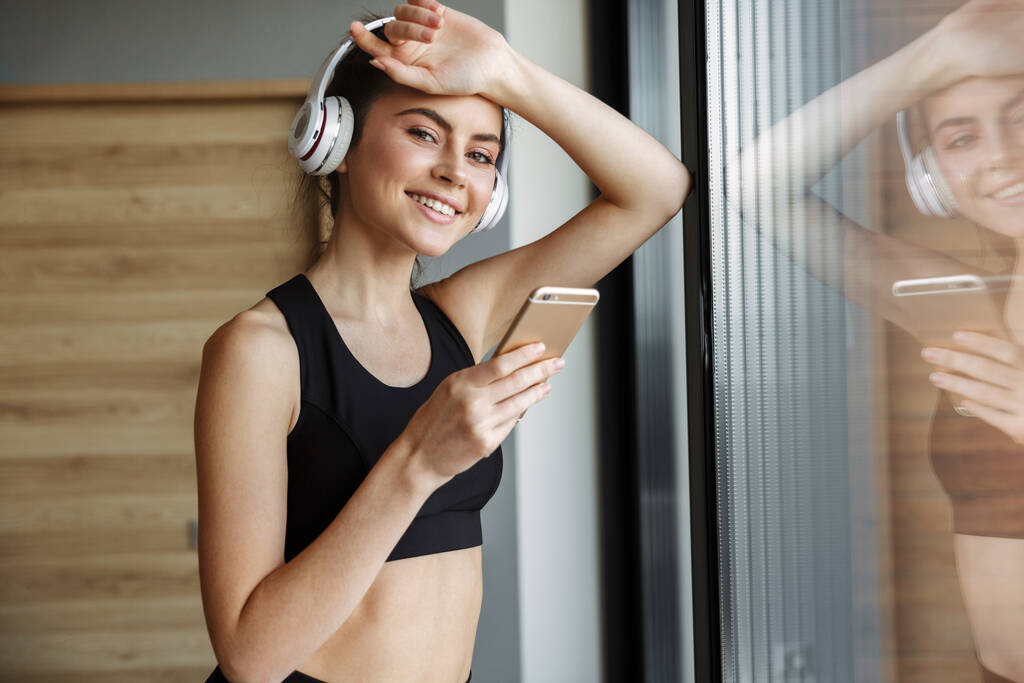 Photo of smiling seductive woman in sportswear using headphones and smartphone while leaning on window - Photo, Image
