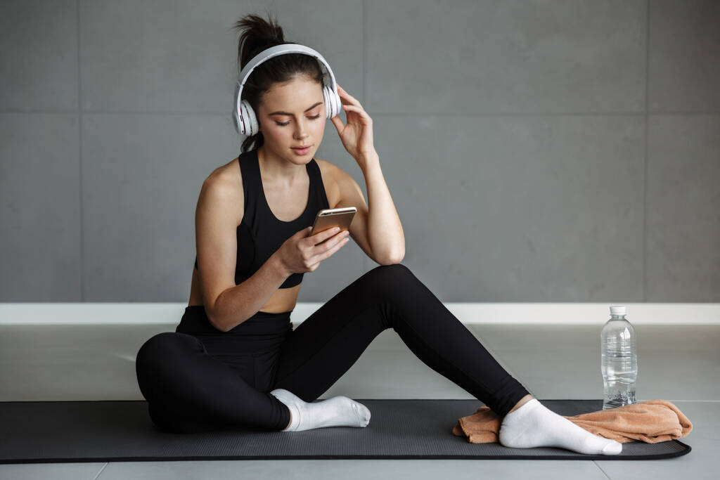 Zdjęcie miłej uwodzicielki w odzieży sportowej używającej słuchawek i telefonu komórkowego podczas siedzenia na macie - Zdjęcie, obraz