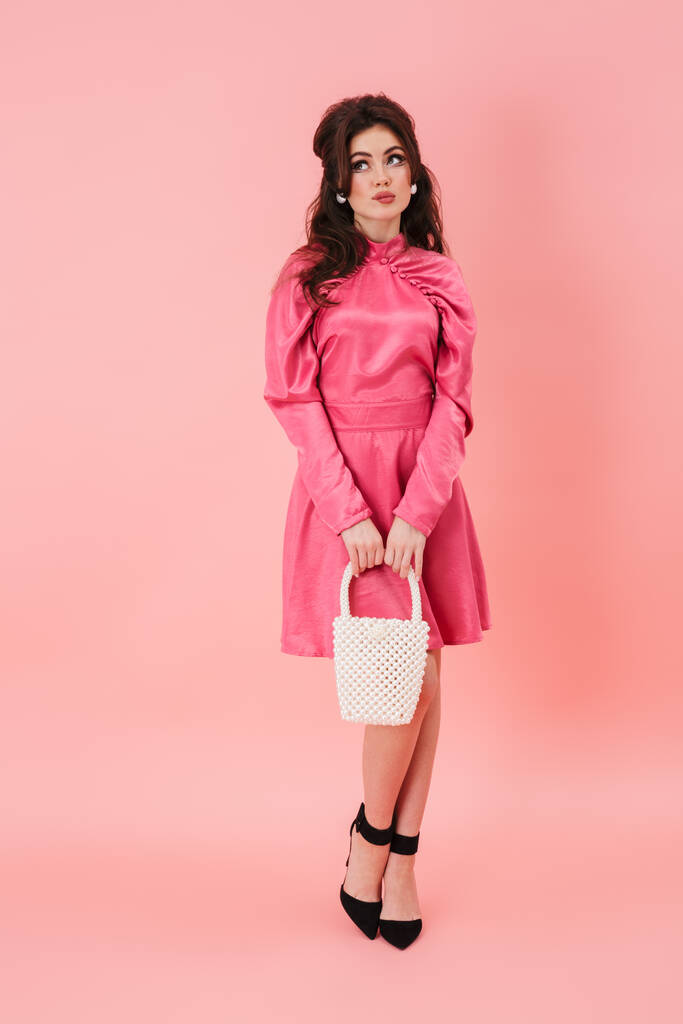 Полный портрет красивой молодой брюнетки с ярким макияжем в модной одежде, стоящей изолированно на розовом фоне, держа кошелек, глядя в сторону
 - Фото, изображение