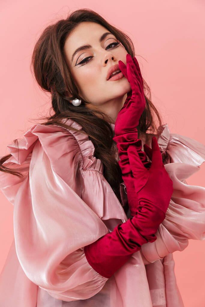 Πορτρέτο μιας όμορφης νεαρής μελαχρινής γυναίκας με φωτεινό μακιγιάζ που φοράει μοντέρνα ρούχα και στέκεται απομονωμένη σε ροζ φόντο, ποζάροντας - Φωτογραφία, εικόνα