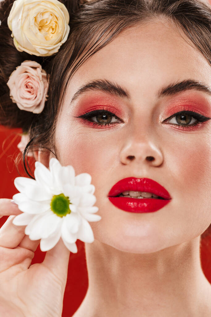 Όμορφη νεαρή μελαχρινή γυναίκα με ένα καταπληκτικό floral χτένισμα φορώντας μακιγιάζ ποζάρουν με χαμομήλι απομονώνονται σε κόκκινο φόντο - Φωτογραφία, εικόνα