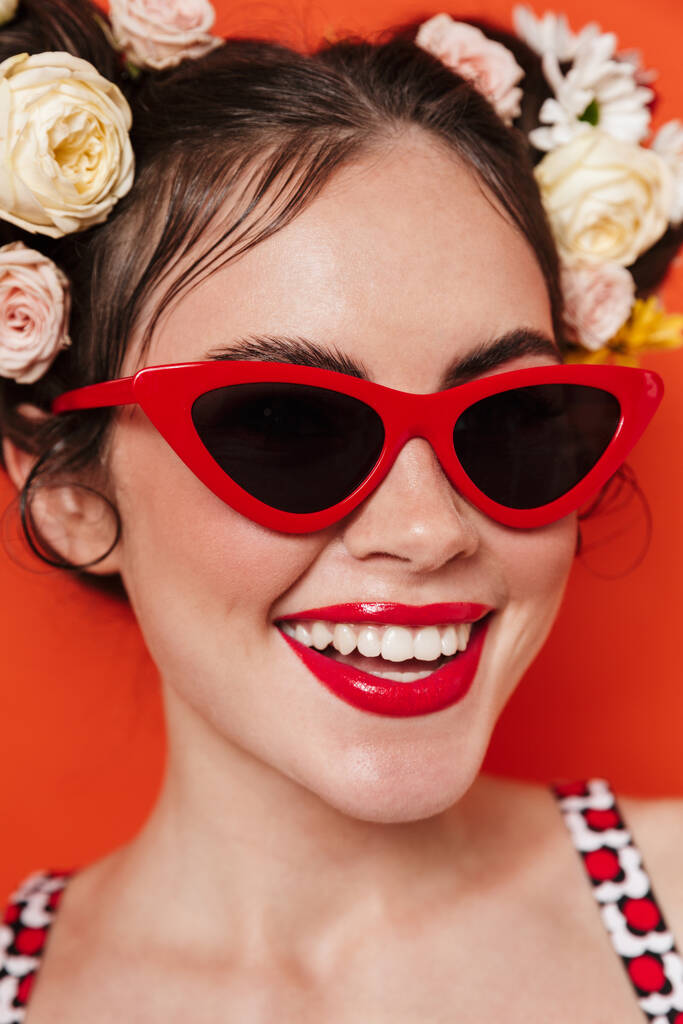 Close up portrait of a beautiful young brunette woman with a floral hairstyle and bright makeup posing isolated over red background, wearing sunglasses - Photo, Image