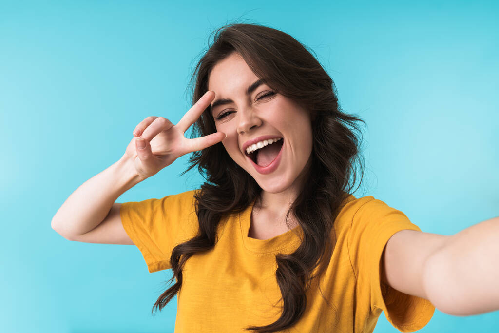 Image of happy positive young pretty woman posing isolated over blue wall background take a selfie by camera showing peace gesture. - Photo, Image